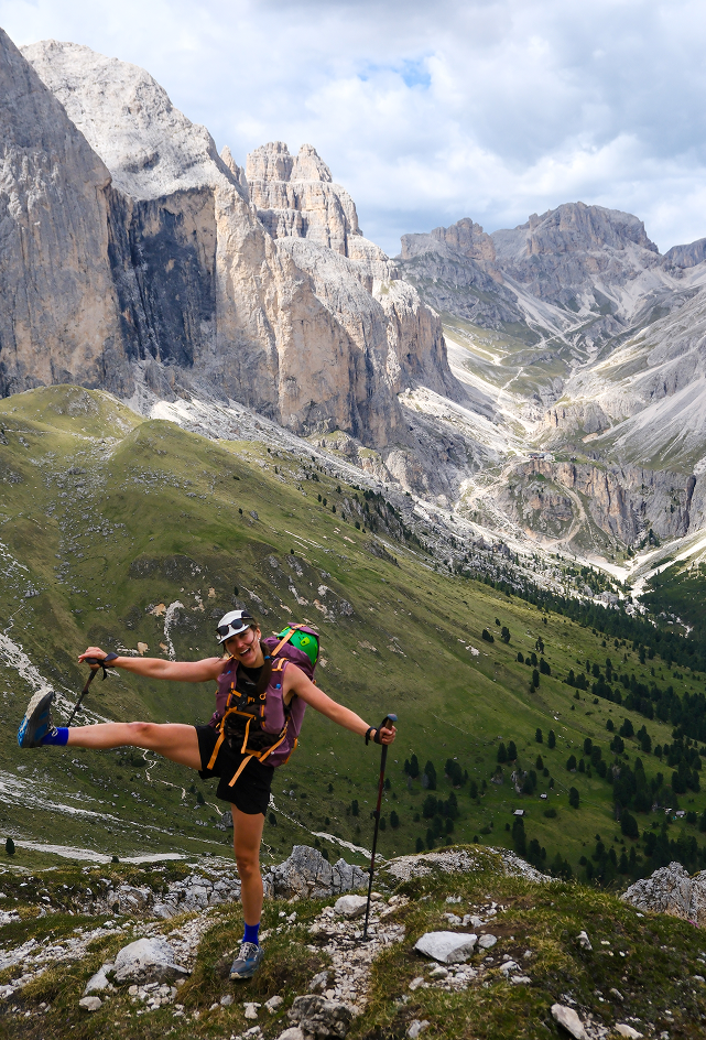 Laura balancing playfully on one leg with trekking poles on a rocky alpine summit, a loaded backpack and climbing helmet behind her